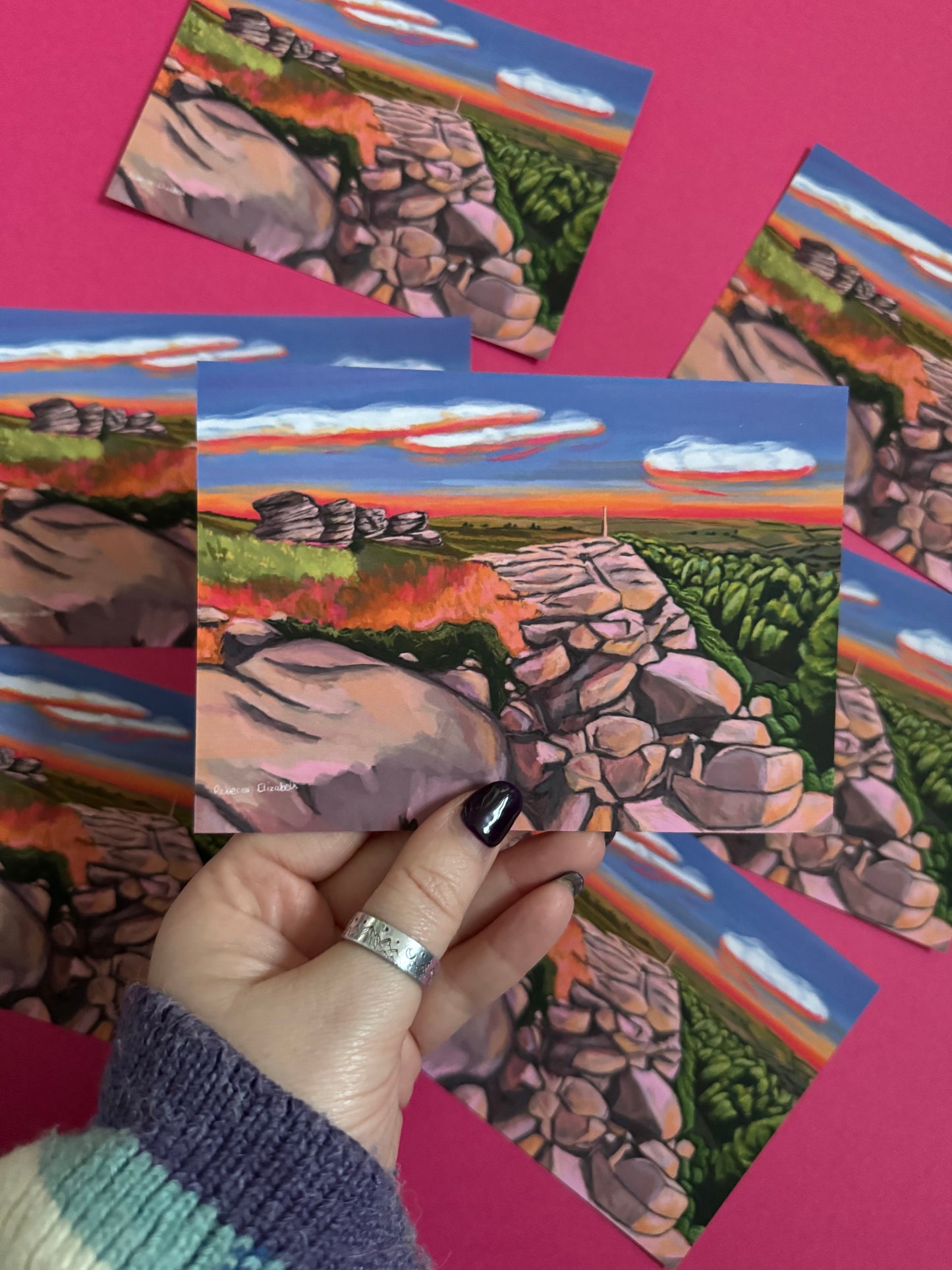 An illustrated postcard of Birchen Edge in the Peak District. With the rock face, rock outcrops named after ships from the battle of Trafalgar and Nelsons Monument all set against a sunset sky with blue, yellow, orange, and pink glow.