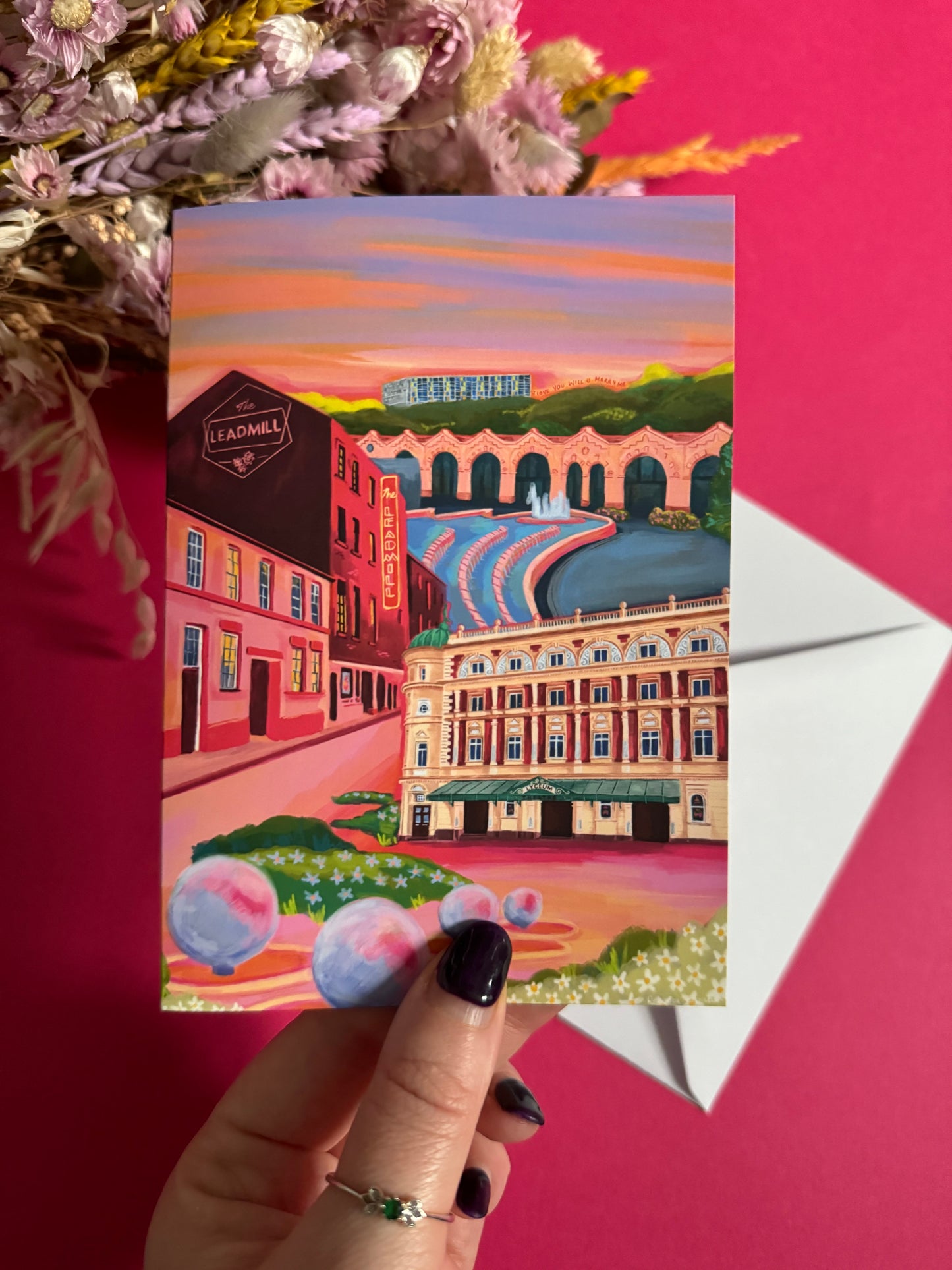 An illustrated greeting card of Sheffield city, Yorkshire. Featuring The Leadmill, the Lyceum, Sheffield train station, Sheffield Park Hill flats, park hill graffiti and water features. This illustration is very vibrant with bright pinks and oranges, the places featured are collaged together against a purple and orange sky. There is a small signature in white the corner. Designed by Rebecca Gibbs at Rebecca Elizabeth Draws.