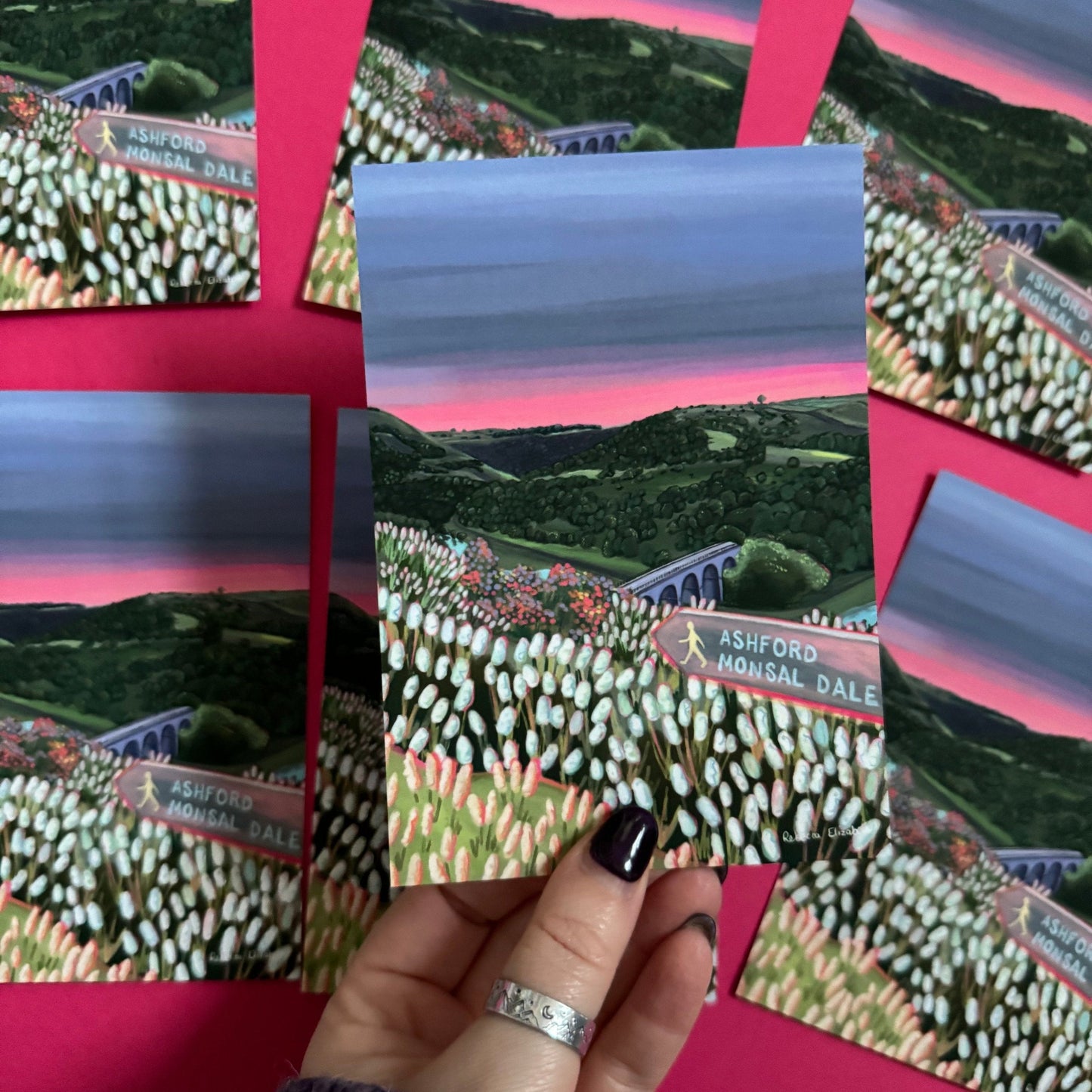 Postcard art of Monsal Head in Derbyshire, Peak District. Rolling green hills filled with trees, an old viaduct over the river Wye and white flowers in the foreground. The sky is a setting sun with pink on the horizon fading into dark blue grey.