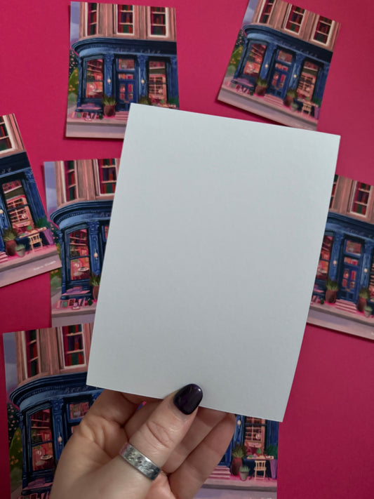 Store front illustration in a deep blue with antique vintage design, inside the book café is a bookcase with jewel tone books, a coffee stand with mugs & cake. The are tables inside and out and upstairs are 3 windows with shelves of books inside. Designed by Rebecca Elizabeth Draws.