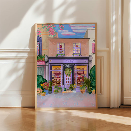 illustration of the bakery, a lilac purple shop front surrounded by older vintage buildings. To the left the window has red polka dot curtains, underneath a pink shop front with daisies on. Above the shop are 2 windows with yellow gingham curtains, a soft brown leather chair & a pink bed with a teddy bear. Inside the bakery the are lots of cakes & cupcakes, bouquets of flowers. Outside the shop there is plant pots full of colourful flowers & sunflowers. The sign has daisies & strawberries on.