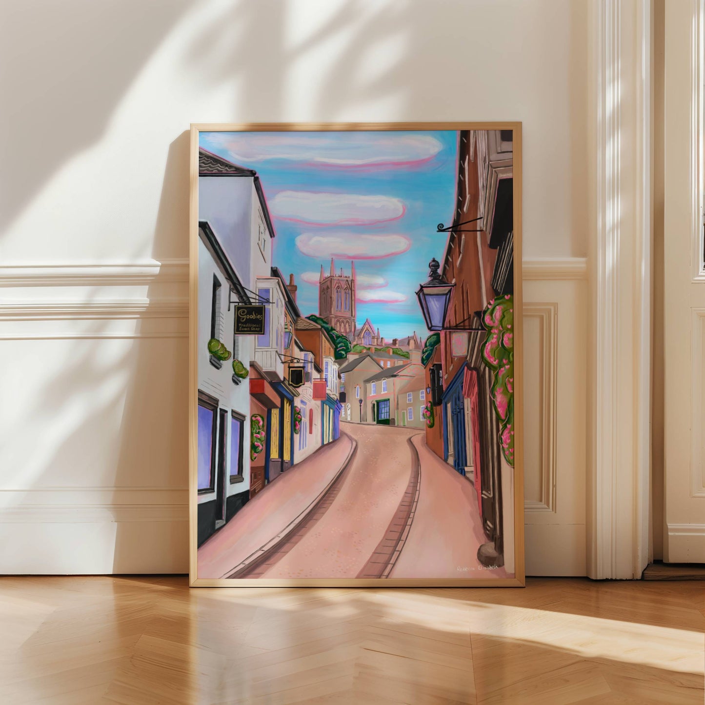 illustration of steep hill in Lincoln, Lincolnshire England UK. IN the background sits Lincoln Cathedral in soft browns & pinks. the sky is a bright blue with fluffy white clouds and a pink highlight. in the foreground the shops that can be found on steep hill, with a mixture of old fashioned buildings and modern signage. The are pink highlights throughout and little pops of greenery with pink flowers.