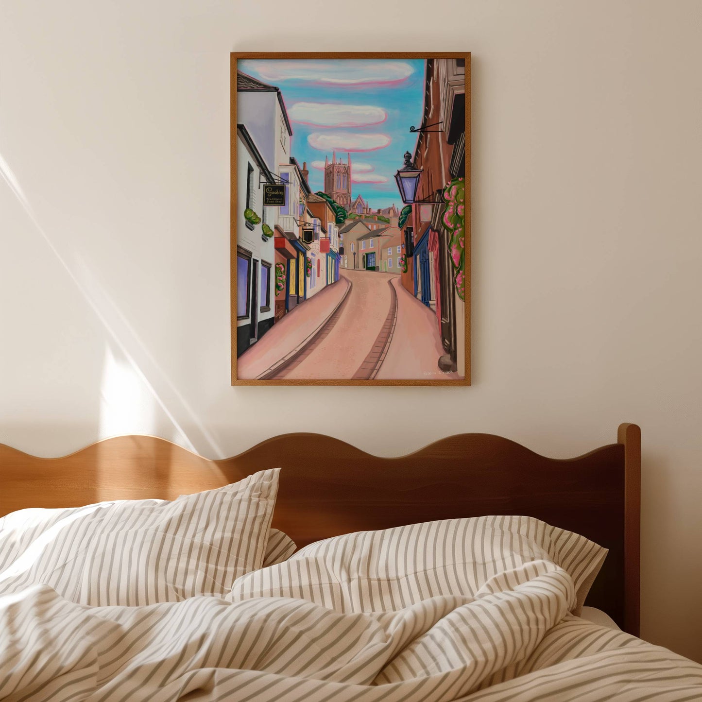 illustration of steep hill in Lincoln, Lincolnshire England UK. IN the background sits Lincoln Cathedral in soft browns & pinks. the sky is a bright blue with fluffy white clouds and a pink highlight. in the foreground the shops that can be found on steep hill, with a mixture of old fashioned buildings and modern signage. The are pink highlights throughout and little pops of greenery with pink flowers.