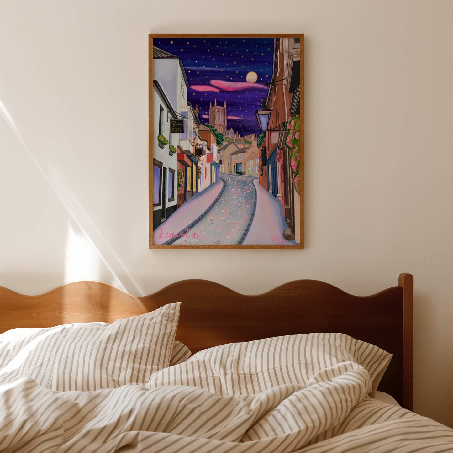 illustration of steep hill in Lincoln, Lincolnshire England UK. IN the background sits Lincoln Cathedral in soft browns & pinks. the sky is a night sky in deep blue with pink cloud tendrils & glittering stars & moon. in the foreground the shops that can be found on steep hill, with a mixture of old fashioned buildings and modern signage. The are pink highlights throughout and little pops of greenery with pink flowers.