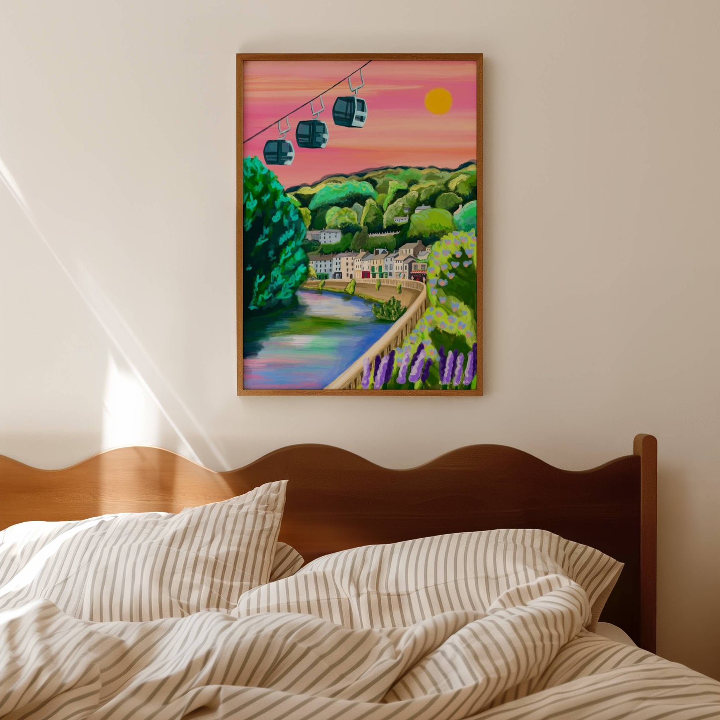 A vibrant and colourful illustration of the beautiful Derbyshire Dales village, Matlock Bath. This illustration features the famous cable cars in the top left hand corner. Then a look across from the river, with the houses and shops set against the big hills. Lots of greenery and a bright pink sky with a bright shining sun. There is a small signature in white the corner. Designed by Rebecca Gibbs at Rebecca Elizabeth Draws.