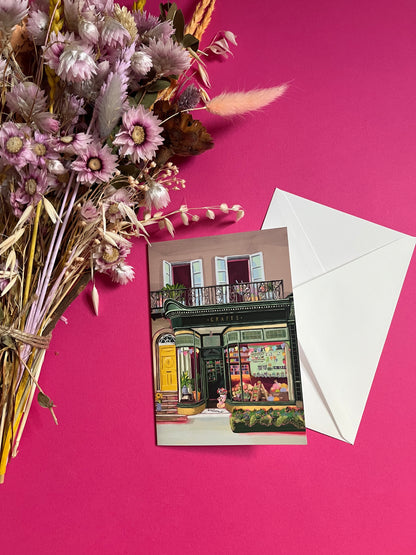 A colourful illustration of a craft shop with the store front in green and looking through the window and at all the goodies. Sewing, drawing, painting, we have it all here at the craft shop. On a greeting card with a green back & logo.