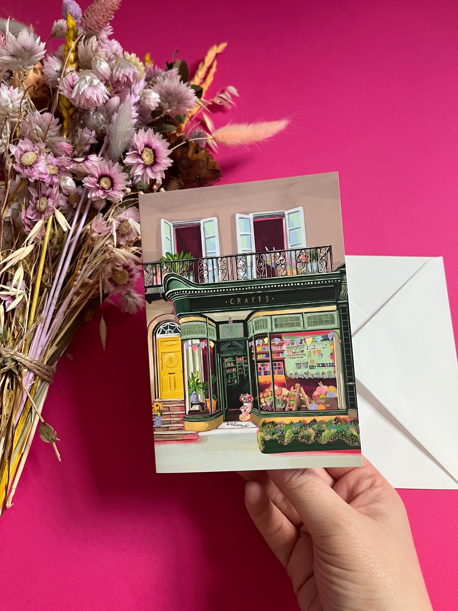 A colourful illustration of a craft shop with the store front in green and looking through the window and at all the goodies. Sewing, drawing, painting, we have it all here at the craft shop. On a greeting card with a green back & logo.