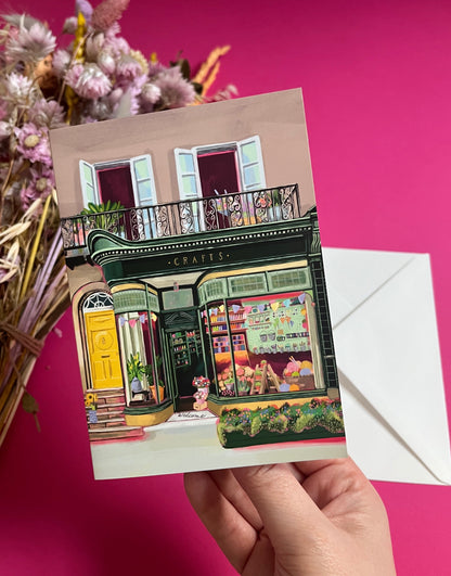 A colourful illustration of a craft shop with the store front in green and looking through the window and at all the goodies. Sewing, drawing, painting, we have it all here at the craft shop. On a greeting card with a green back & logo.