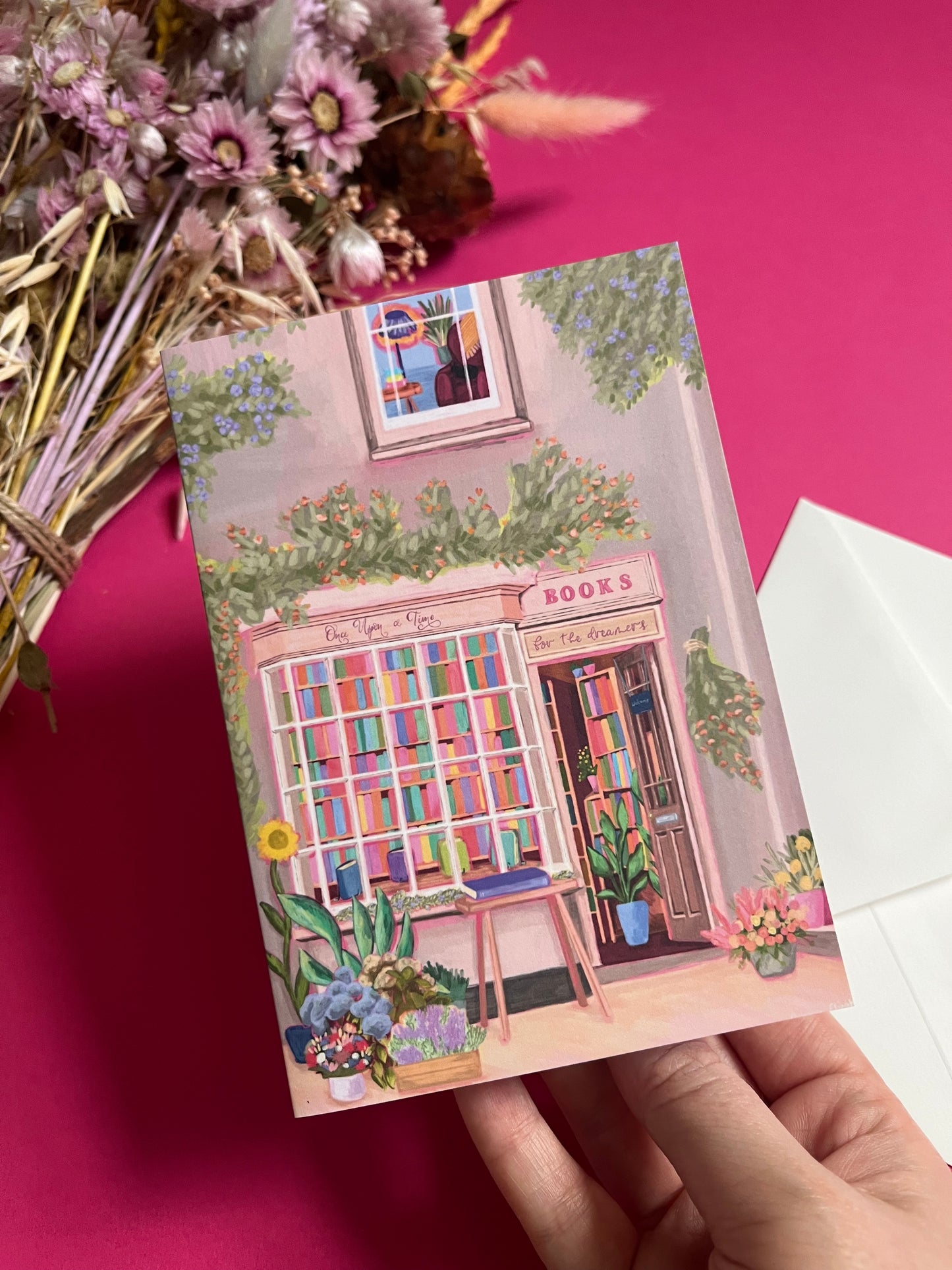 An illustration of a cute whimsical bookshop, the window shows the bookshelves inside full of bright coloured book spines with lots of flowers. The back of the card is pink with the Rebecca Elizabeth Draws logo. 