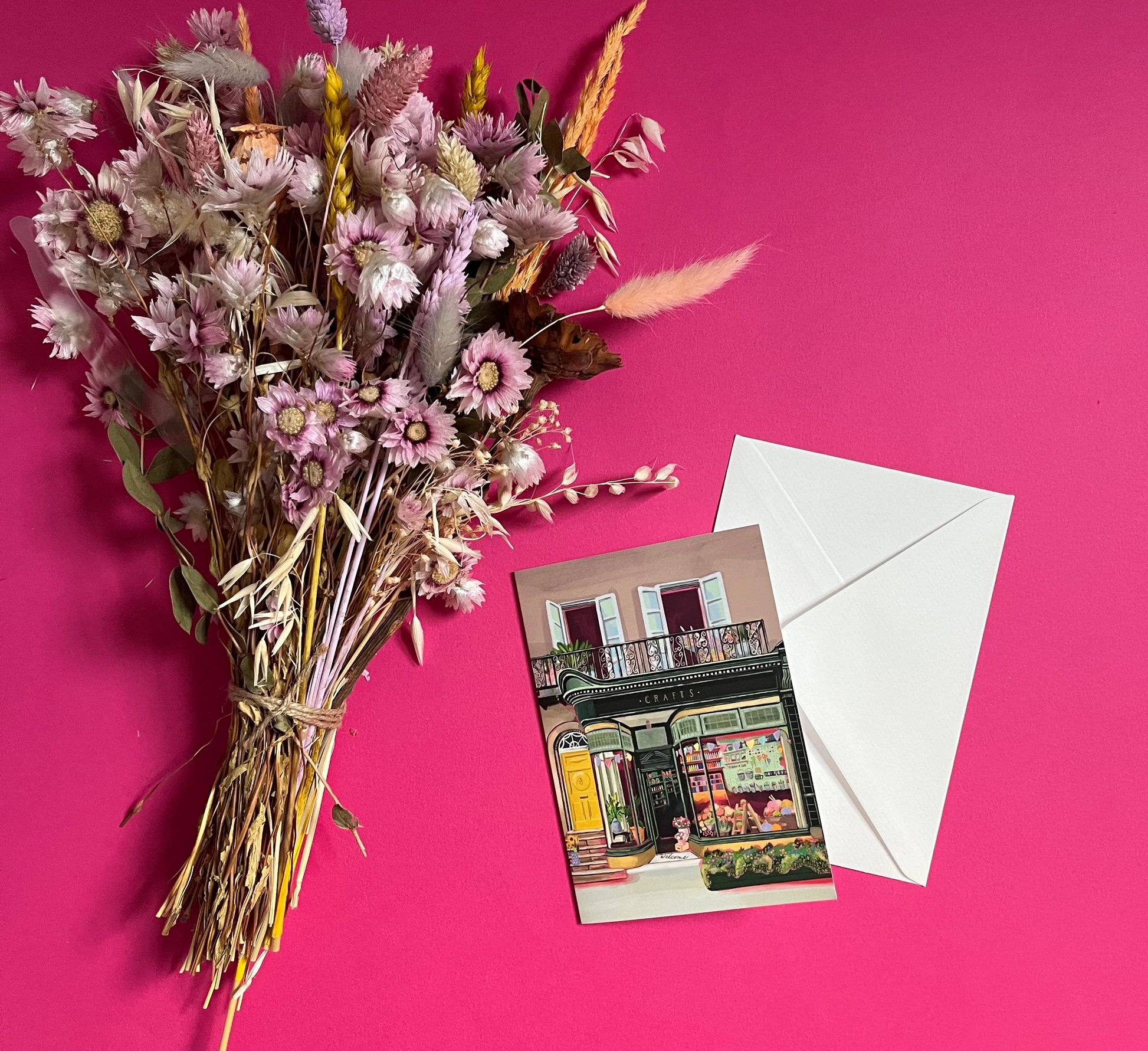 A colourful illustration of a craft shop with the store front in green and looking through the window and at all the goodies. Sewing, drawing, painting, we have it all here at the craft shop. On a greeting card with a green back & logo.