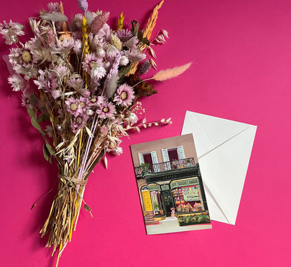 A colourful illustration of a craft shop with the store front in green and looking through the window and at all the goodies. Sewing, drawing, painting, we have it all here at the craft shop. On a greeting card with a green back & logo.