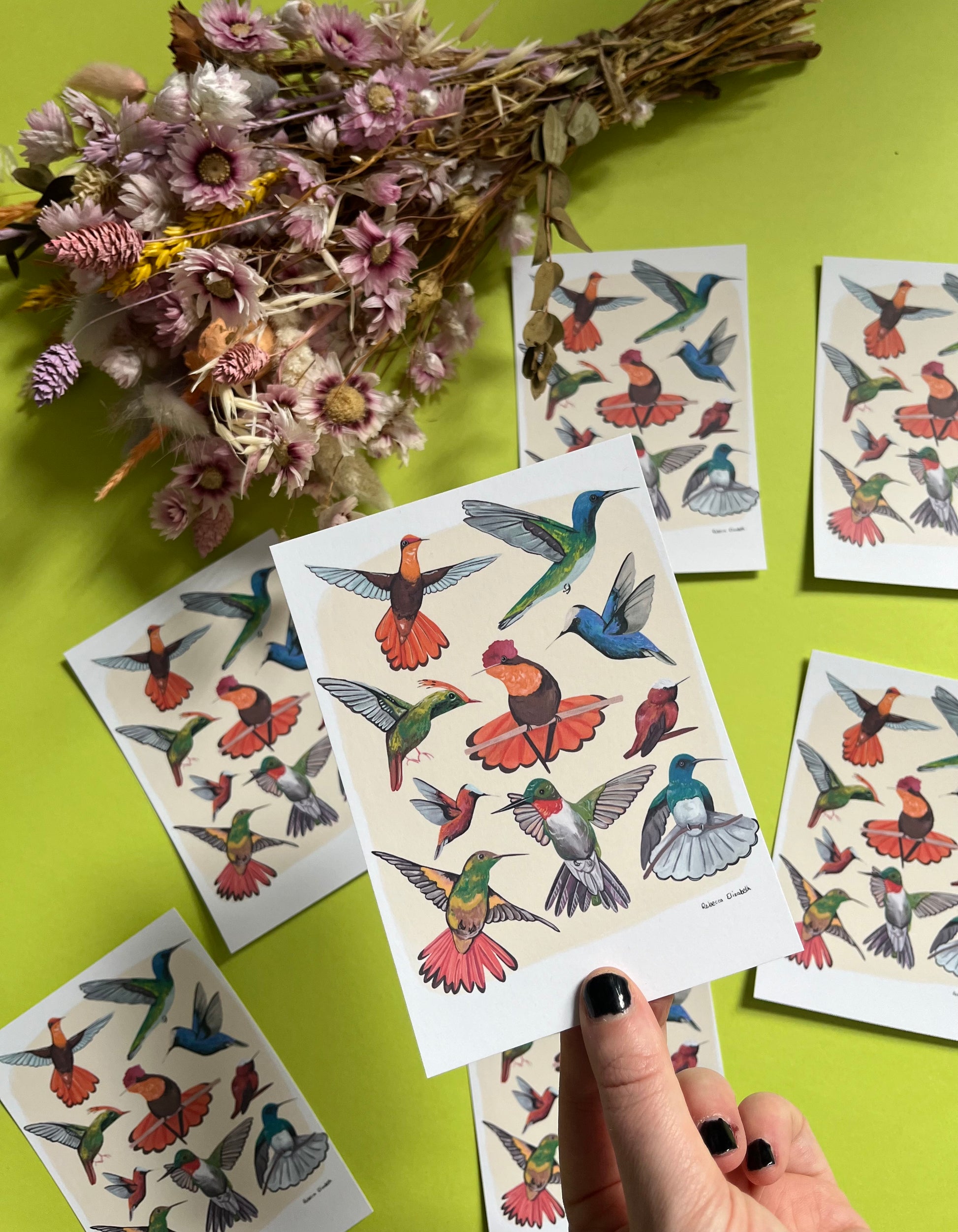 Hummingbirds hand painted in gouache and edited together digitally on a cream background with a small signature featured in the right hand corner. These small birds are green, blue, red, orange and yellow. Designed by Rebecca Gibbs on a postcard at Rebecca Elizabeth Draws.