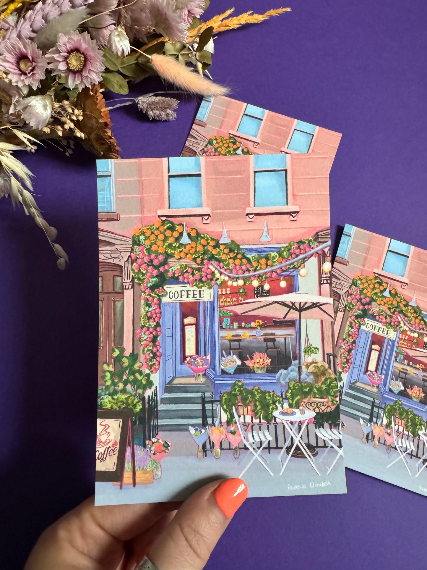 A coffee shop illustration with a blue exterior, an outside table and umbrella and building windows surrounding. This illustration is filled with lots of plants and flowers and features cups of coffee and baked goods. Illustrated by Rebecca Elizabeth Draws. 