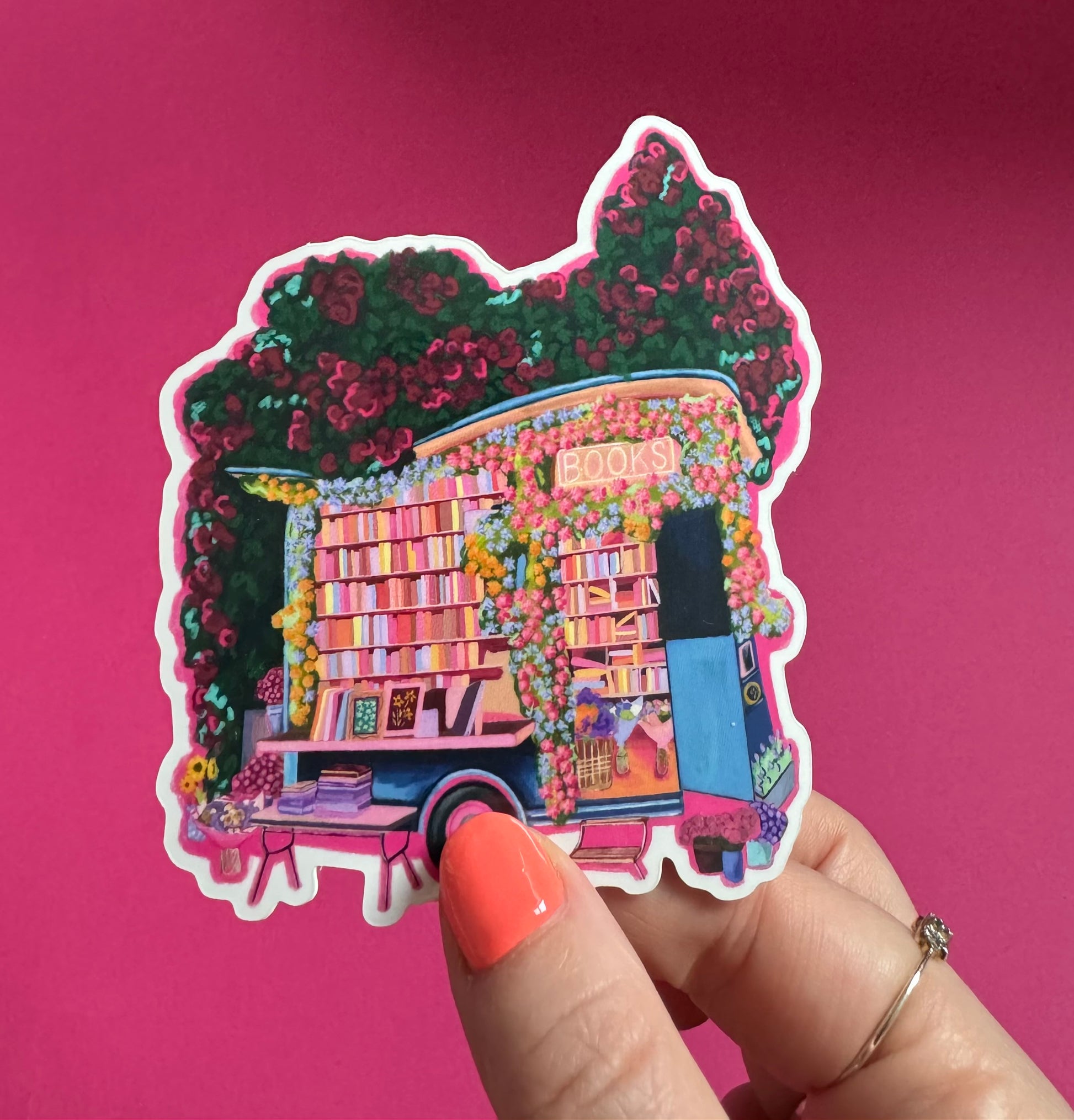 Illustration of a van opened up showing lots of bookshelves stacked with books, art frames out front and lots of blossoming pink, orange and blue flowers over the top. A big tree with pink roses over hangs and behind sits old fashioned buildings with quaint architecture and balconies. A whimsical fantasy sticker.