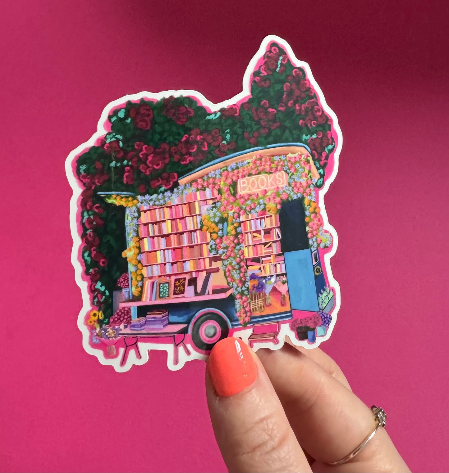 Illustration of a van opened up showing lots of bookshelves stacked with books, art frames out front and lots of blossoming pink, orange and blue flowers over the top. A big tree with pink roses over hangs and behind sits old fashioned buildings with quaint architecture and balconies. A whimsical fantasy sticker.