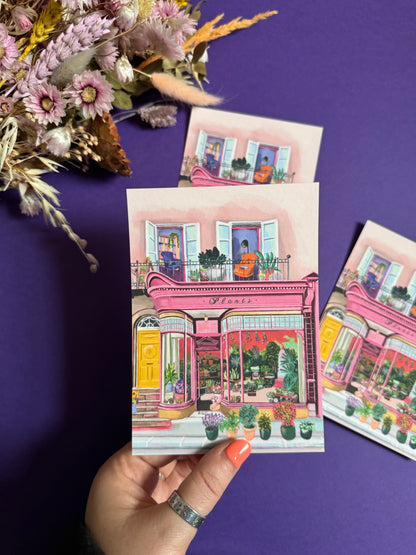 Pink shop front of a plant store with lots of greenery inside, a balcony above with windows showing chairs and a bookcase and more flowers. A yellow door to the side with a sunflower vase resting on the steps by Rebecca Elizabeth Draws. 