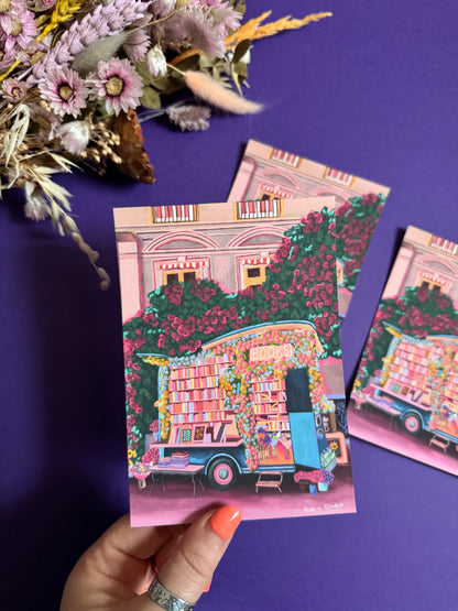 Illustration of a van opened up showing lots of bookshelves stacked with books, art frames out front and lots of blossoming pink, orange and blue flowers over the top. A big tree with pink roses over hangs and behind sits old fashioned buildings with quaint architecture and balconies. A whimsical fantasy print. 