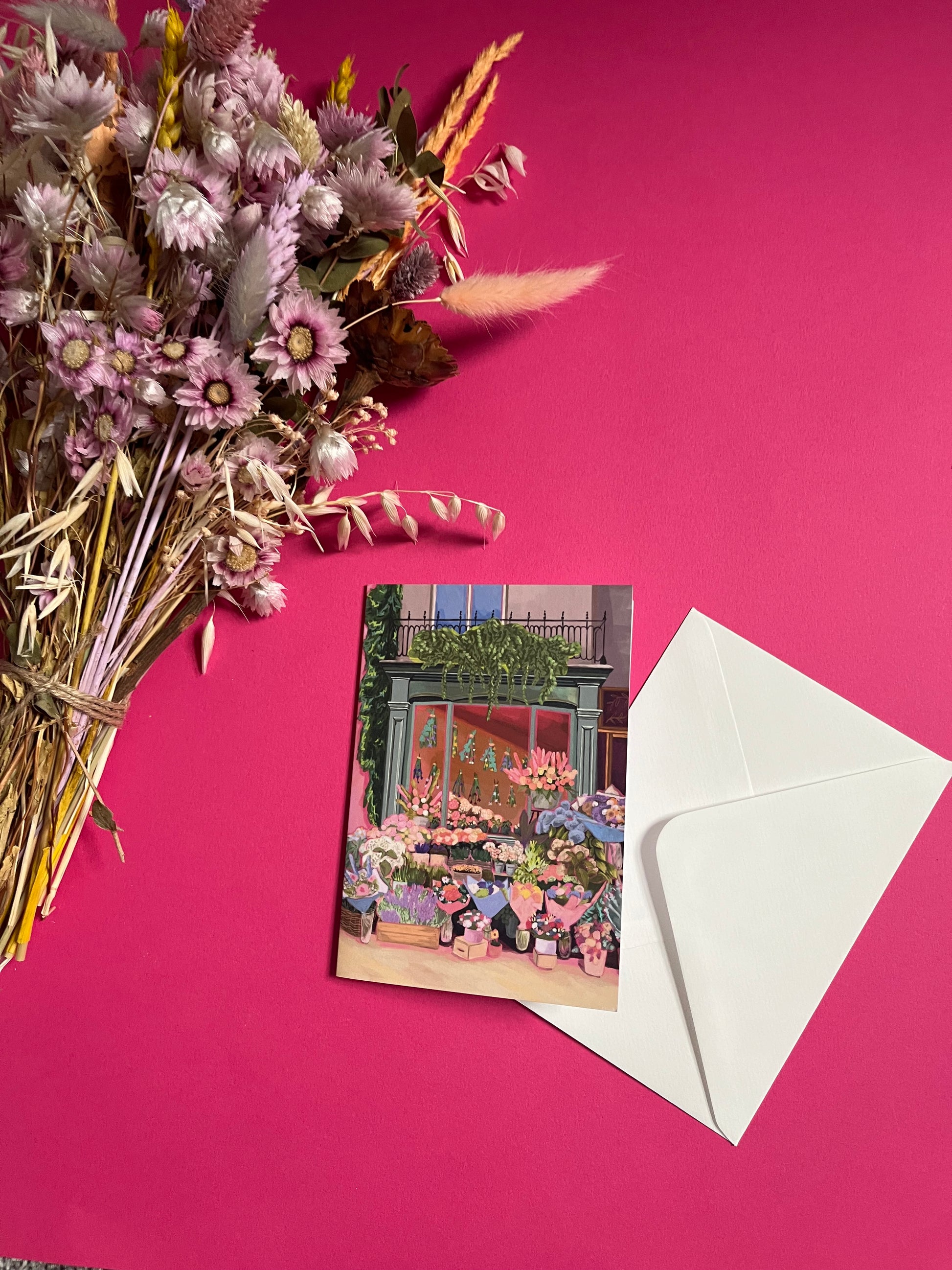 Greeting card of a beautiful flower shop, the window shows the flowers drying inside and outside there are rows of bunches of flowers and pots of plants. Soft brown and pink with bright highlights throughout. With a pink back piece & logo.