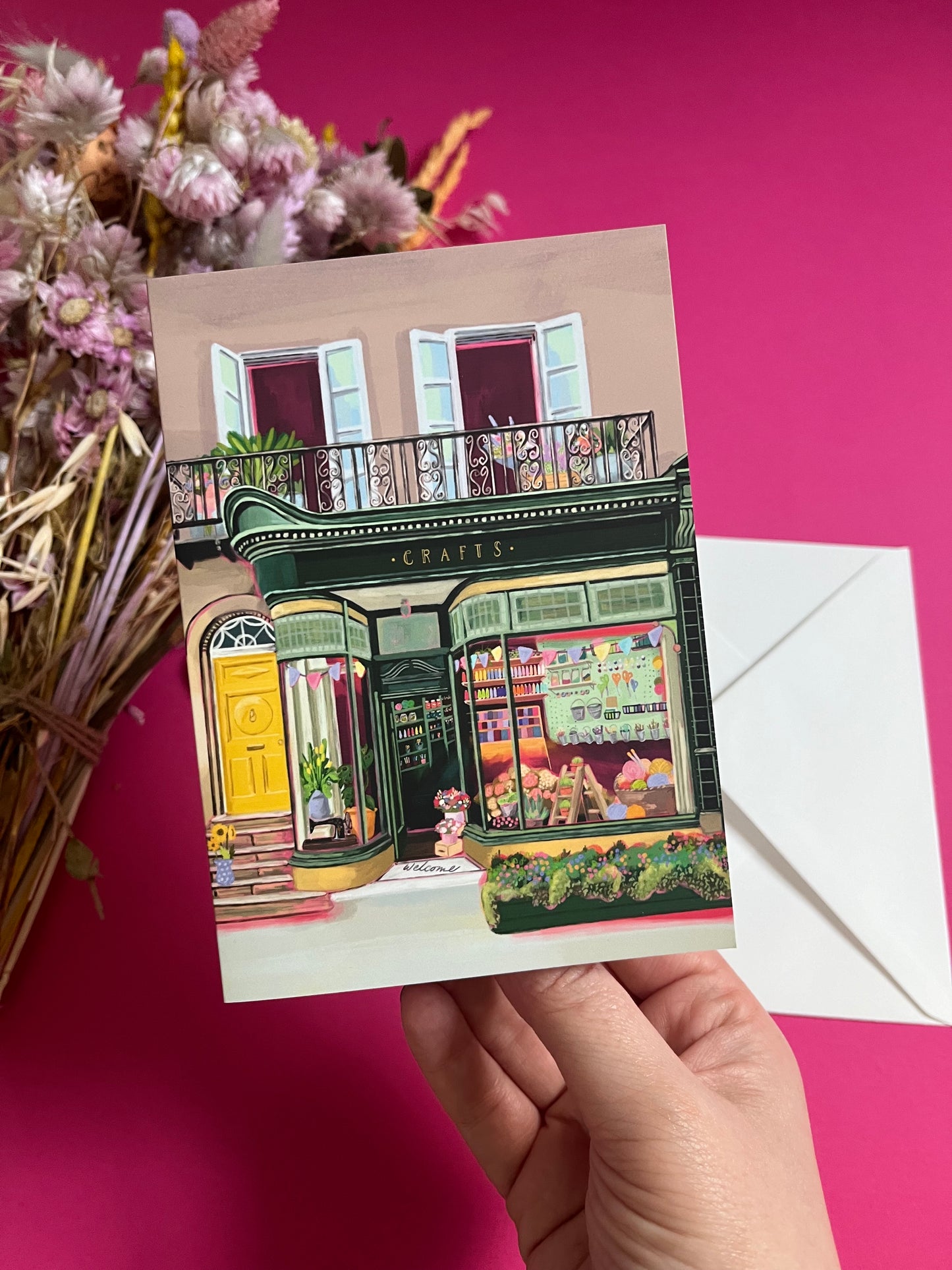 A colourful illustration of a craft shop with the store front in green and looking through the window and at all the goodies. Sewing, drawing, painting, we have it all here at the craft shop. On a greeting card with a green back & logo.