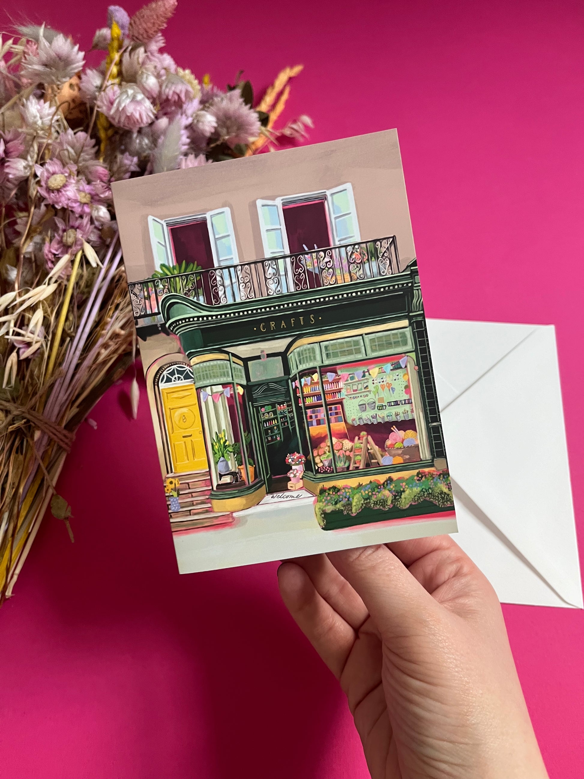 A colourful illustration of a craft shop with the store front in green and looking through the window and at all the goodies. Sewing, drawing, painting, we have it all here at the craft shop. On a greeting card with a green back & logo.