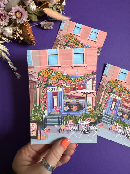 A coffee shop illustration with a blue exterior, an outside table and umbrella and building windows surrounding. This illustration is filled with lots of plants and flowers and features cups of coffee and baked goods. Illustrated by Rebecca Elizabeth Draws. 