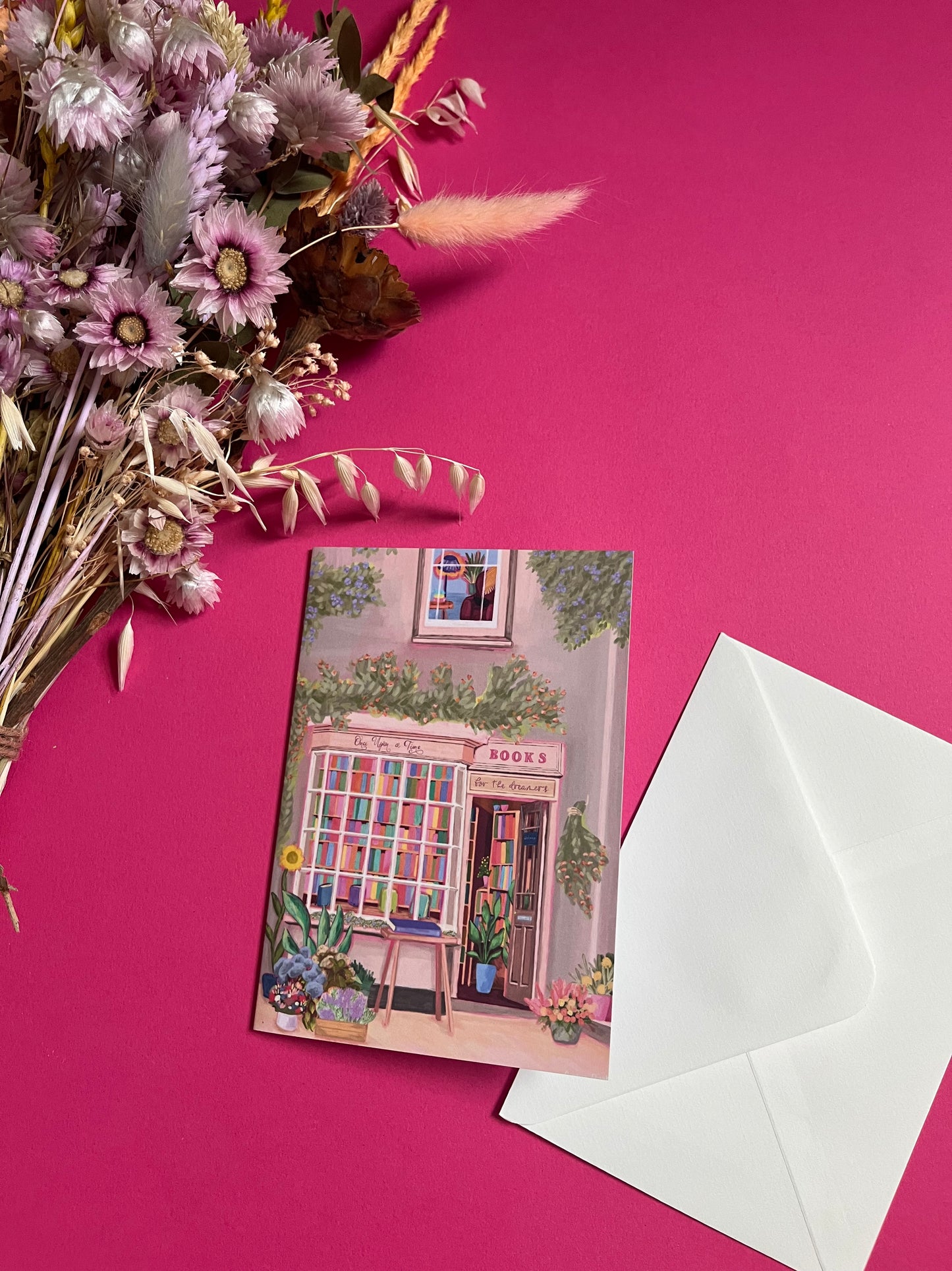 An illustration of a cute whimsical bookshop, the window shows the bookshelves inside full of bright coloured book spines with lots of flowers. The back of the card is pink with the Rebecca Elizabeth Draws logo. 