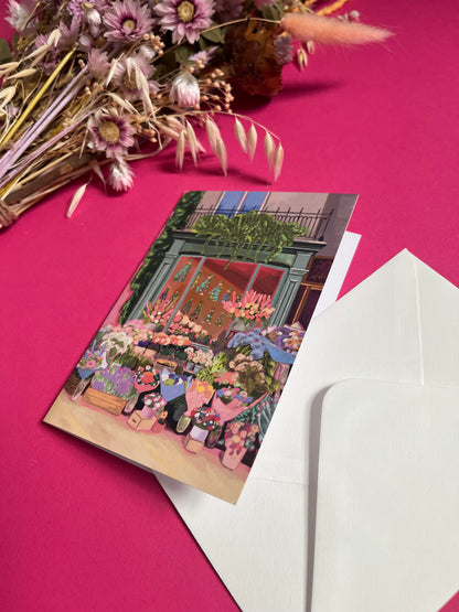 Greeting card of a beautiful flower shop, the window shows the flowers drying inside and outside there are rows of bunches of flowers and pots of plants. Soft brown and pink with bright highlights throughout. With a pink back piece & logo.