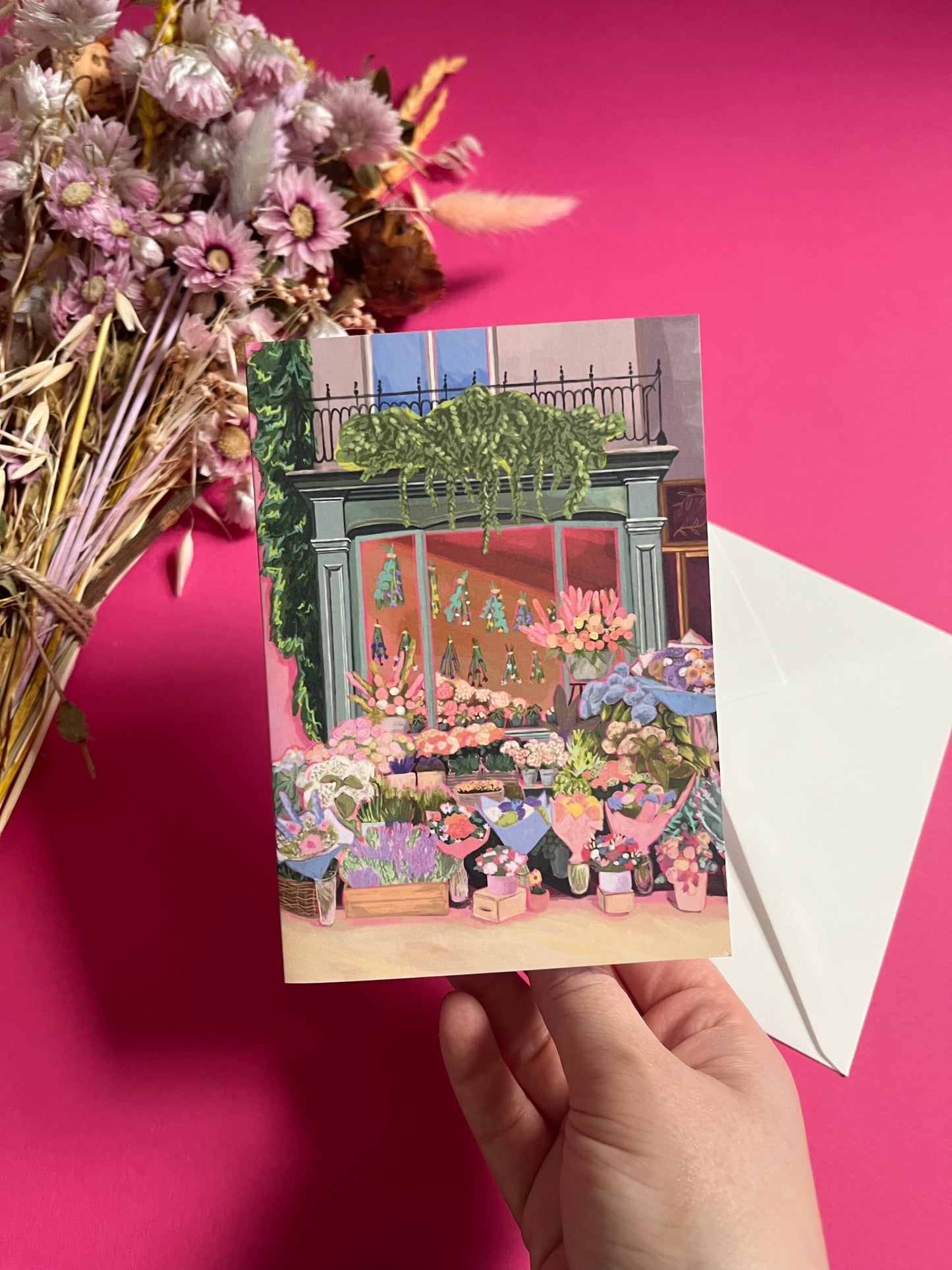 Greeting card of a beautiful flower shop, the window shows the flowers drying inside and outside there are rows of bunches of flowers and pots of plants. Soft brown and pink with bright highlights throughout. With a pink back piece & logo.