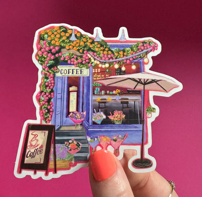 A coffee shop art sticker with a blue exterior, an outside table and umbrella and building windows surrounding. This sticker is filled with lots of plants and flowers & features cups of coffee & baked goods. Illustrated by Rebecca Elizabeth Draws. 