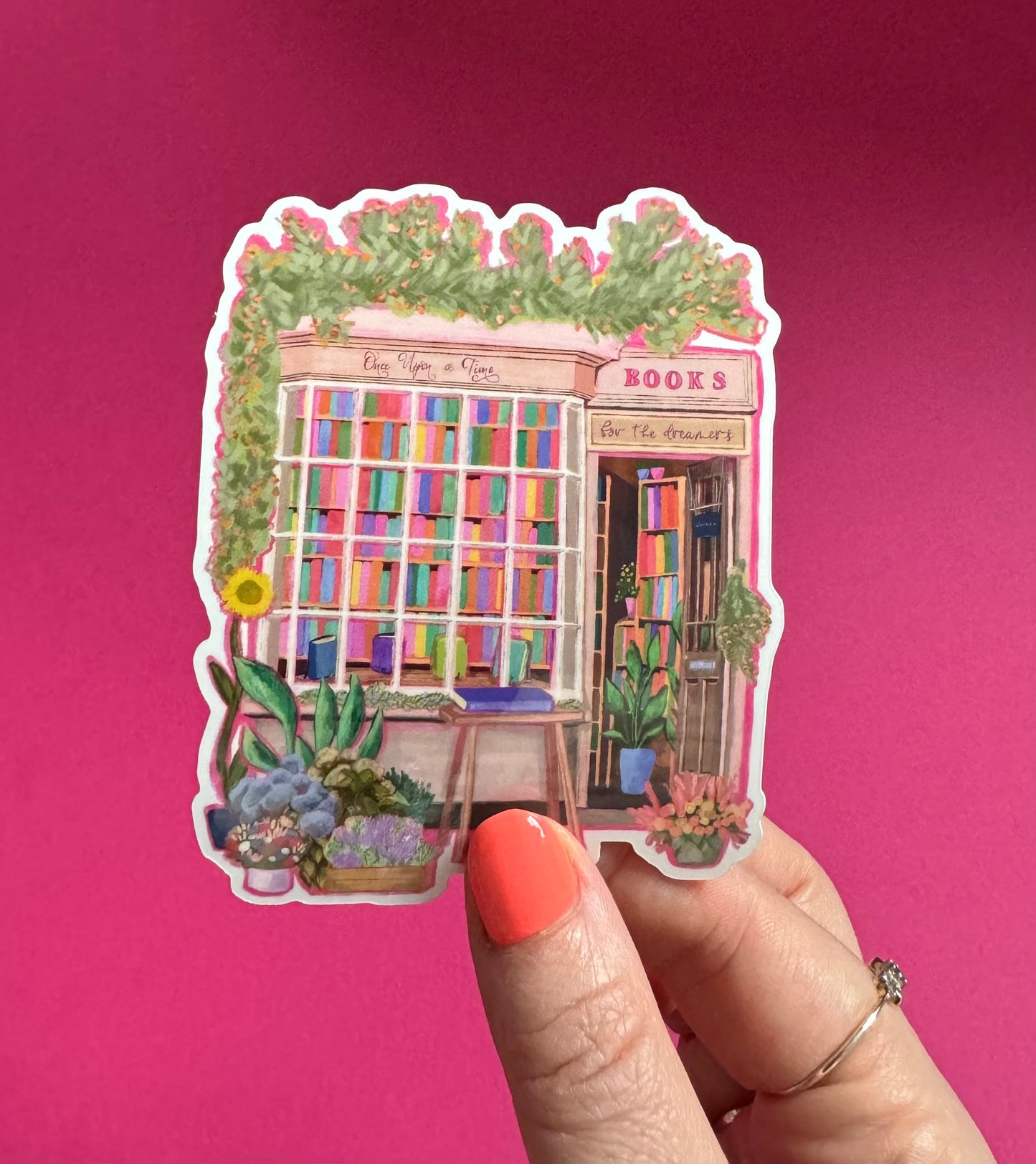 A sticker illustration of a cute bookshop, the window shows the bookshelves inside full of bright coloured book spines. There are books placed stood up at the front. Outside the building is a soft colour with lots of greenery in soft pale greens and orange and lilac flowers. In front of the building there are plant pots and flowers. Art sticker designed by Rebecca Gibbs at Rebecca Elizabeth Draws.