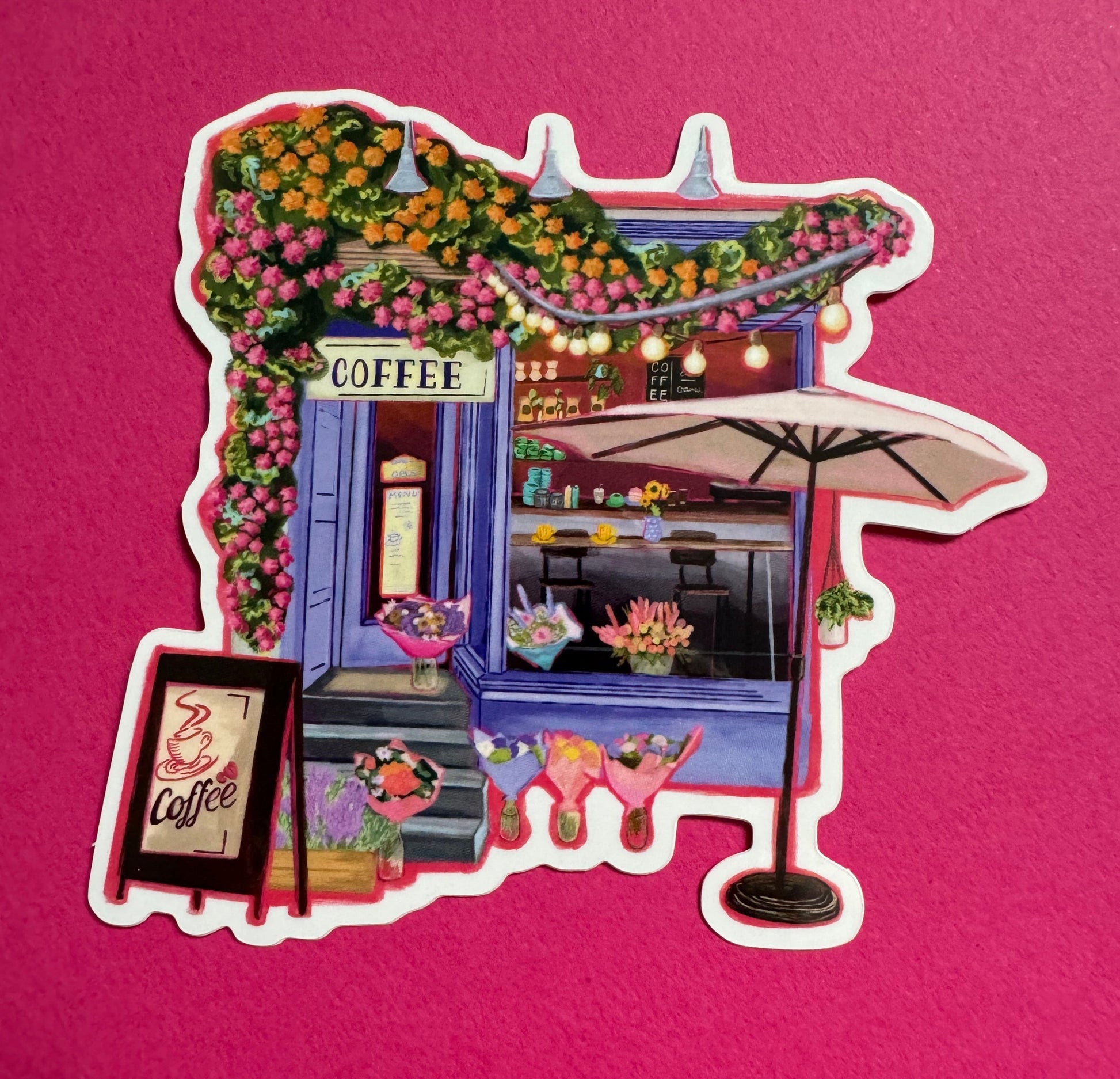 A coffee shop art sticker with a blue exterior, an outside table and umbrella and building windows surrounding. This sticker is filled with lots of plants and flowers & features cups of coffee & baked goods. Illustrated by Rebecca Elizabeth Draws. 