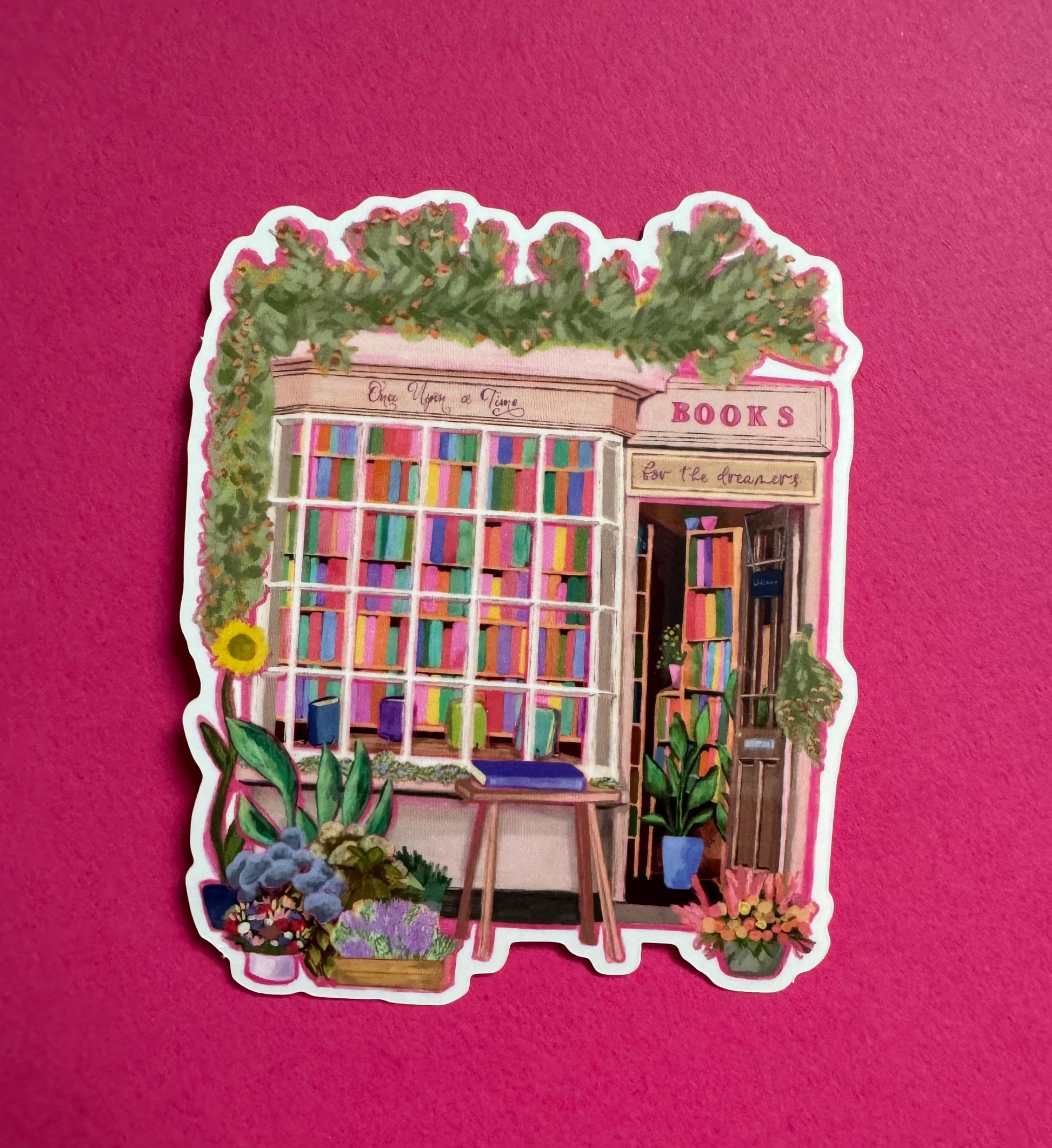 A sticker illustration of a cute bookshop, the window shows the bookshelves inside full of bright coloured book spines. There are books placed stood up at the front. Outside the building is a soft colour with lots of greenery in soft pale greens and orange and lilac flowers. In front of the building there are plant pots and flowers. Art sticker designed by Rebecca Gibbs at Rebecca Elizabeth Draws.