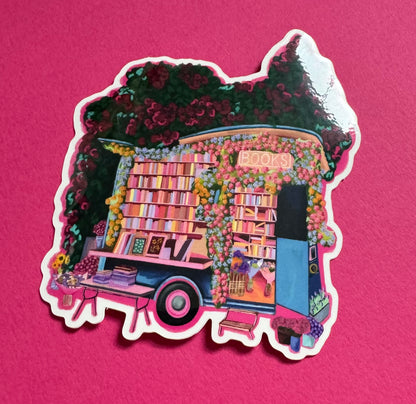 Illustration of a van opened up showing lots of bookshelves stacked with books, art frames out front and lots of blossoming pink, orange and blue flowers over the top. A big tree with pink roses over hangs and behind sits old fashioned buildings with quaint architecture and balconies. A whimsical fantasy sticker.