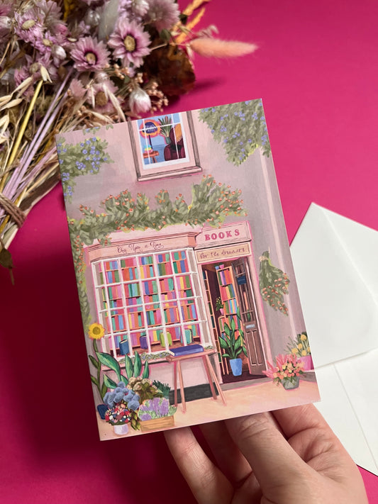 An illustration of a cute whimsical bookshop, the window shows the bookshelves inside full of bright coloured book spines with lots of flowers. The back of the card is pink with the Rebecca Elizabeth Draws logo. 