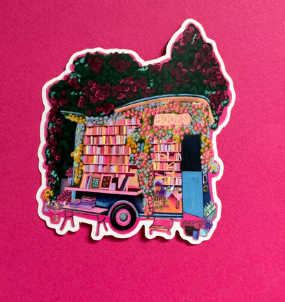 Illustration of a van opened up showing lots of bookshelves stacked with books, art frames out front and lots of blossoming pink, orange and blue flowers over the top. A big tree with pink roses over hangs and behind sits old fashioned buildings with quaint architecture and balconies. A whimsical fantasy sticker.