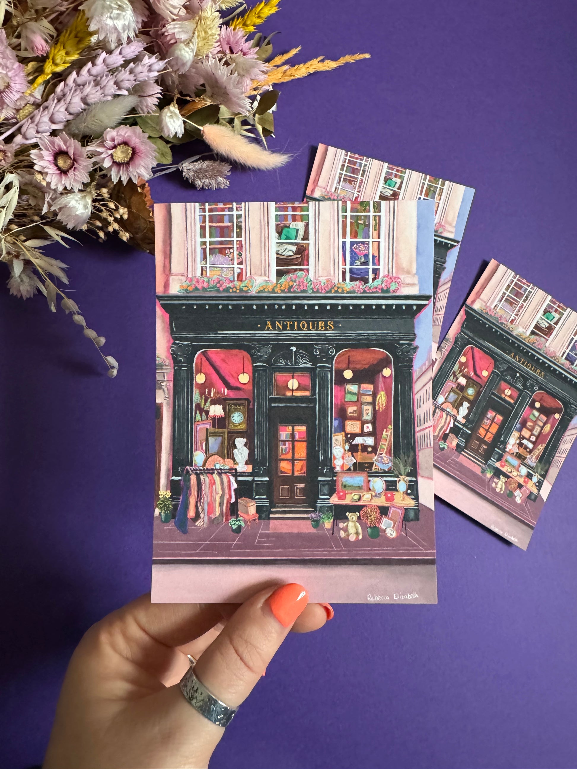 Illustration of a antiques shop front in black with ornate designs, inside is pink and golden with vintage objects such a clocks, statues, picture frames. The illustration gives off dark academia vibes. Designed by Rebecca Elizabeth Draws. 