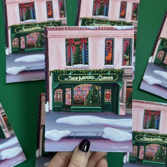 Christmas Illustrated postcard inspired by the film You’ve Got Mail. The Shop Around the Corner is a cosy & whimsical bookshop owned by Kathleen Kelly, inside is lots of fairy lights, Christmas trees, books and baubles. Outside you can see snow and inside the cosy windows with festive decor.  Designed by Rebecca Gibbs at Rebecca Elizabeth Draws.