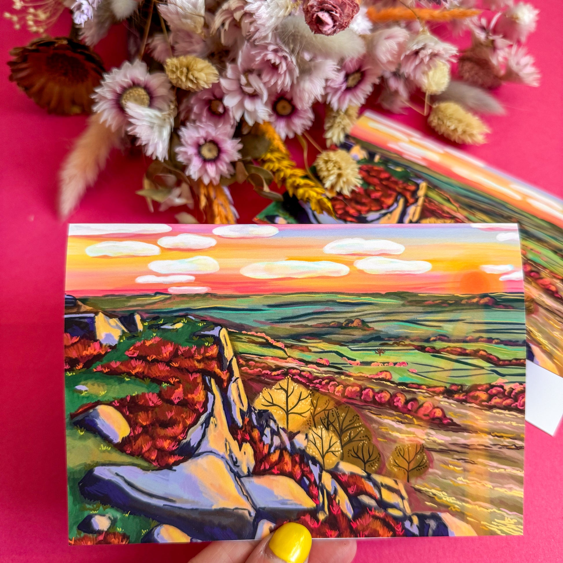Colorful landscape painting held against a pink background with dried flowers.