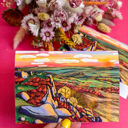 Colorful landscape painting held against a pink background with dried flowers.