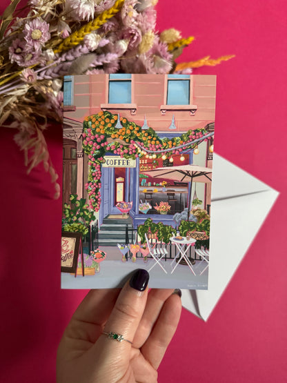 A coffee shop illustrated card with a blue exterior, an outside table and umbrella and building windows surrounding. This illustration is filled with lots of plants and flowers and features cups of coffee and baked goods. Illustrated by Rebecca Elizabeth Draws. 