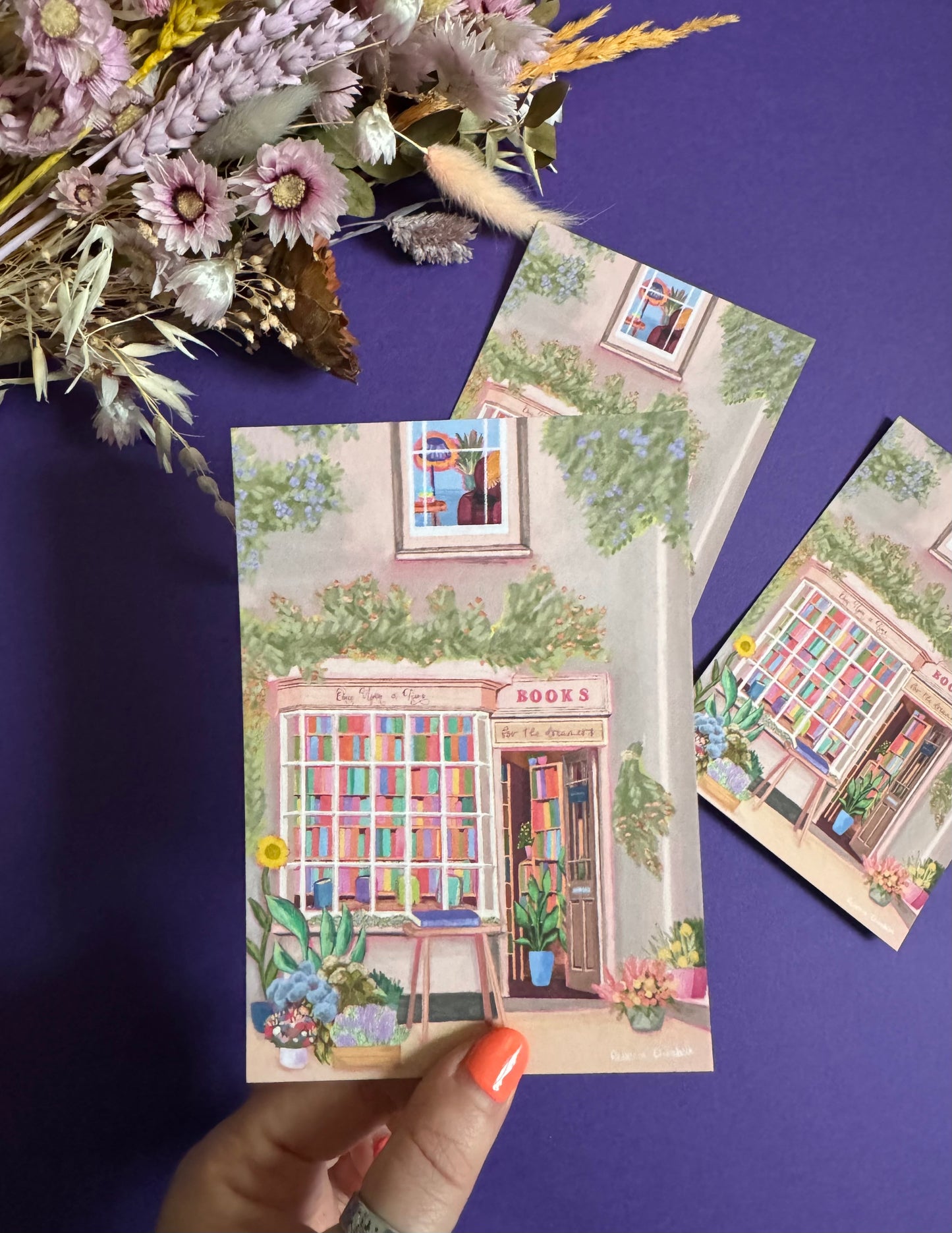 An illustration of a cute bookshop, the window shows the bookshelves inside full of bright coloured book spines. There are books placed stood up at the front. Outside the building is a soft colour with lots of greenery in soft pale greens and orange and lilac flowers. In front of the building there are plant pots and flowers. There is a small signature in white the corner.  Designed by Rebecca Gibbs on a postcard at Rebecca Elizabeth Draws.