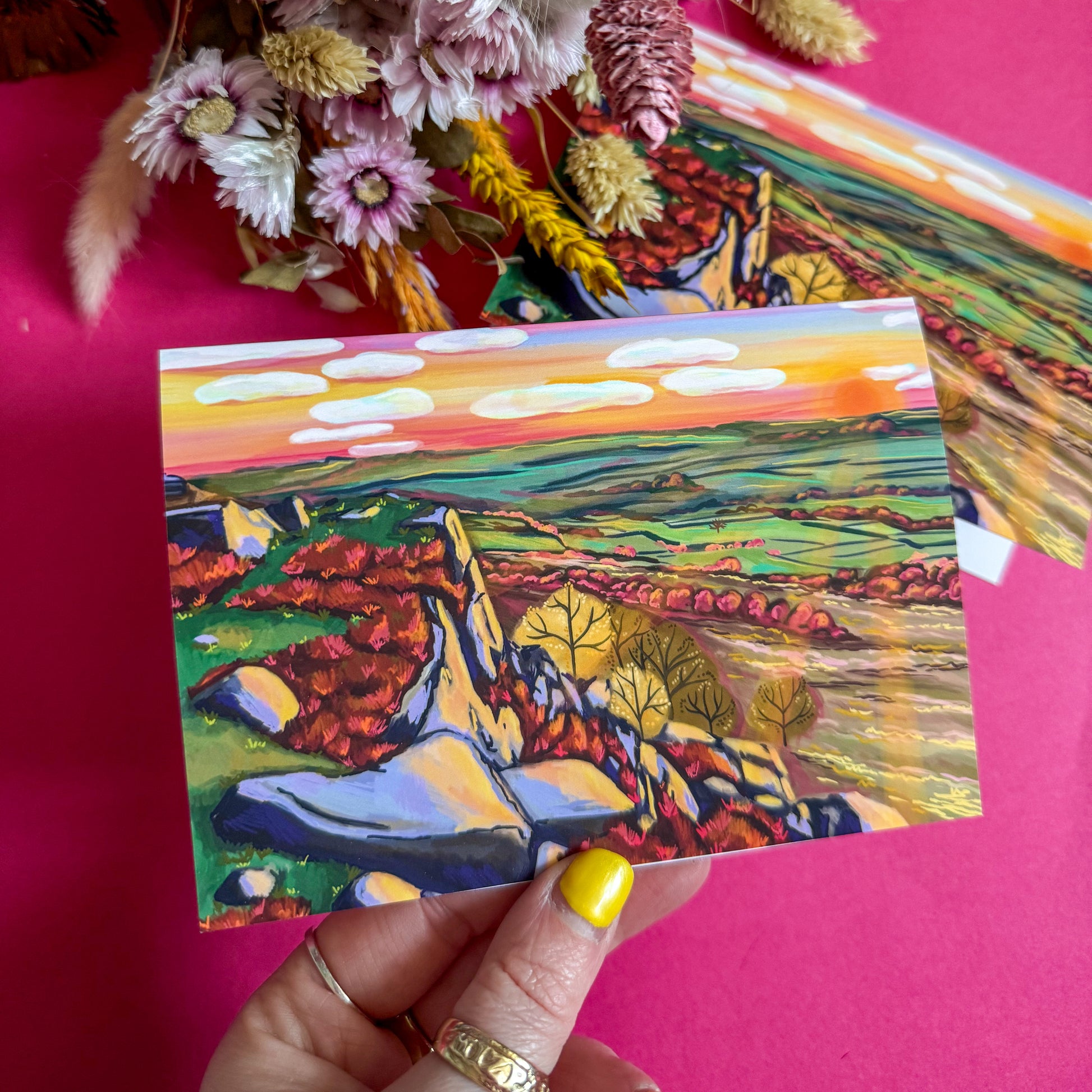 Colorful landscape painting held by a hand with yellow nail polish on a pink background.