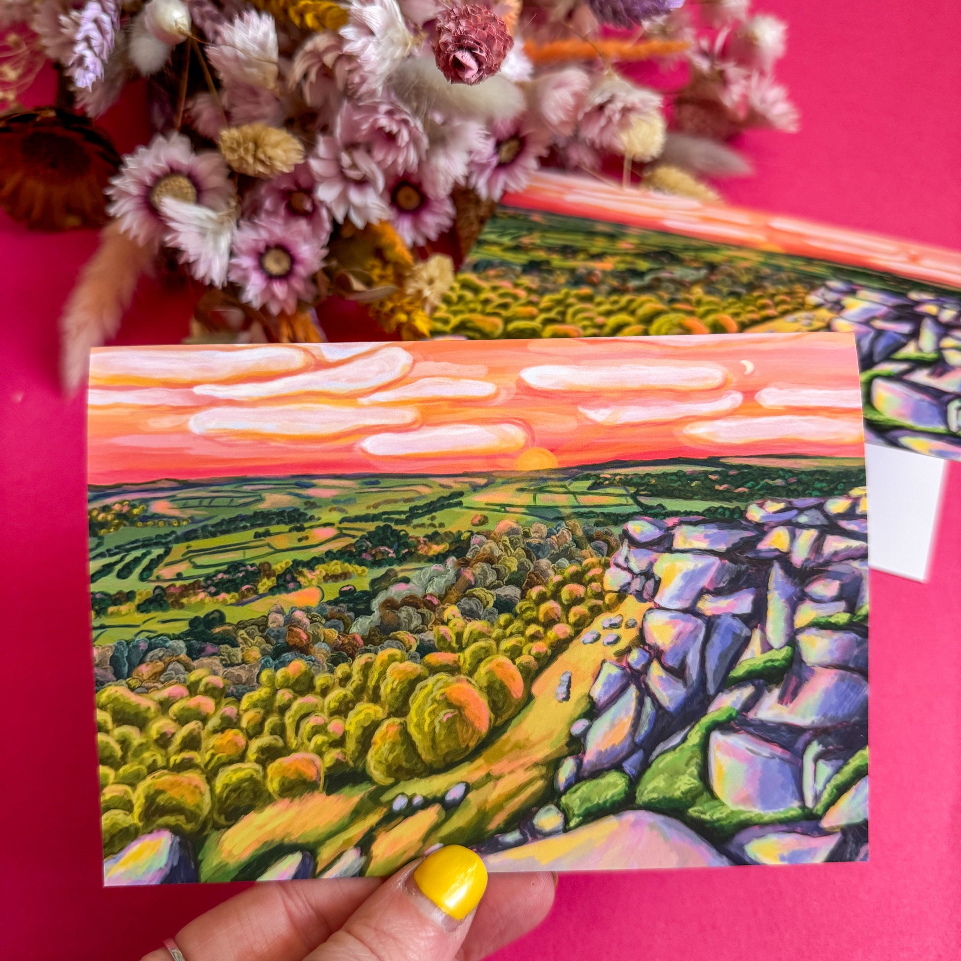 Colorful landscape postcard held by a hand with yellow nail polish against a pink background.