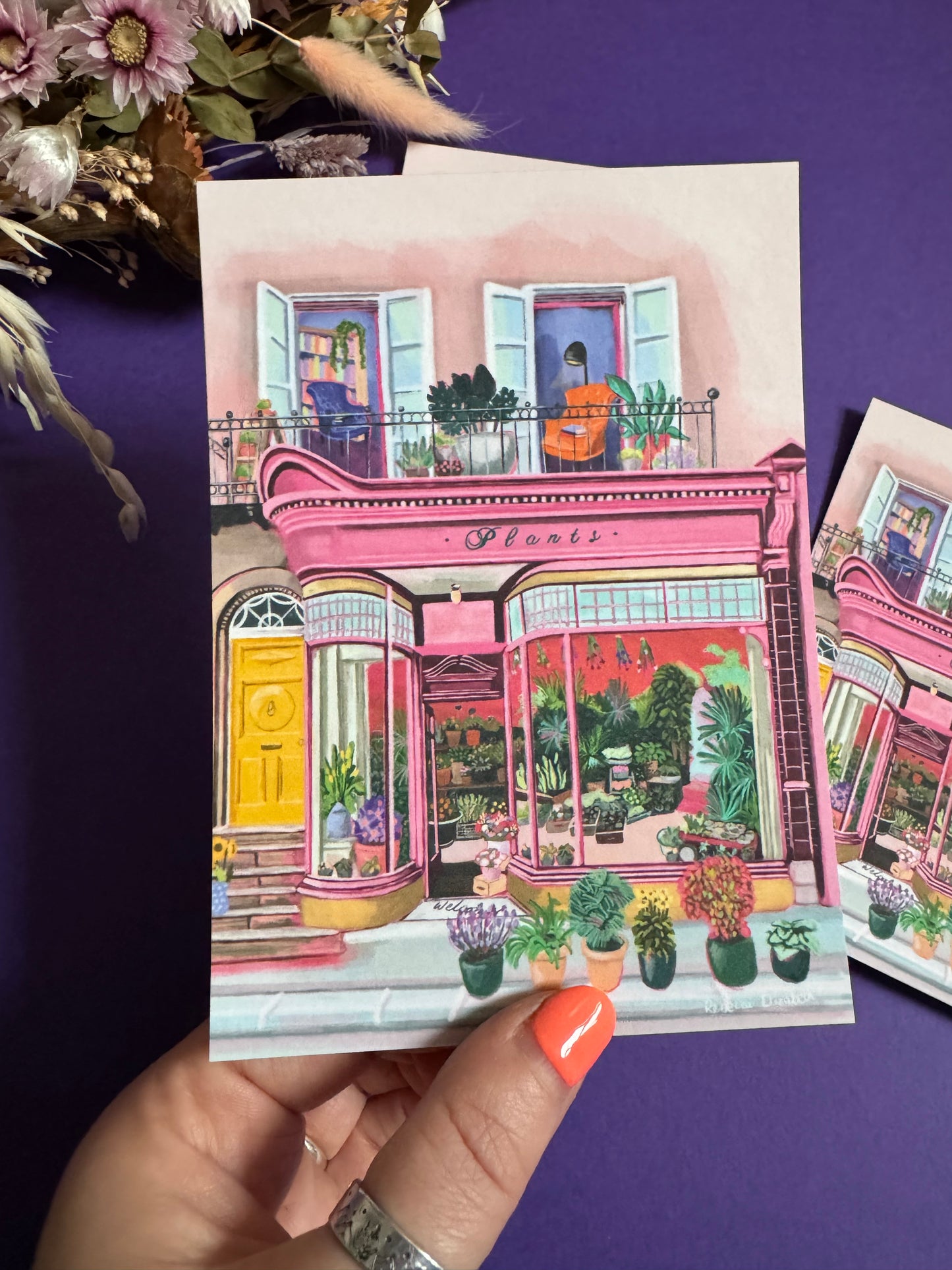 Pink shop front of a plant store with lots of greenery inside, a balcony above with windows showing chairs and a bookcase and more flowers. A yellow door to the side with a sunflower vase resting on the steps by Rebecca Elizabeth Draws. 