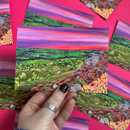A colourful illustration of Curbar edge, Derbyshire with the rock formation to the right covered in pink and golden flowers, yellow flowers in the foreground and then rolling hills to the left with lots of greenery and trees. A stunning bright pink and purple sunset across the sky.  Designed by 
Rebecca Elizabeth draws. 