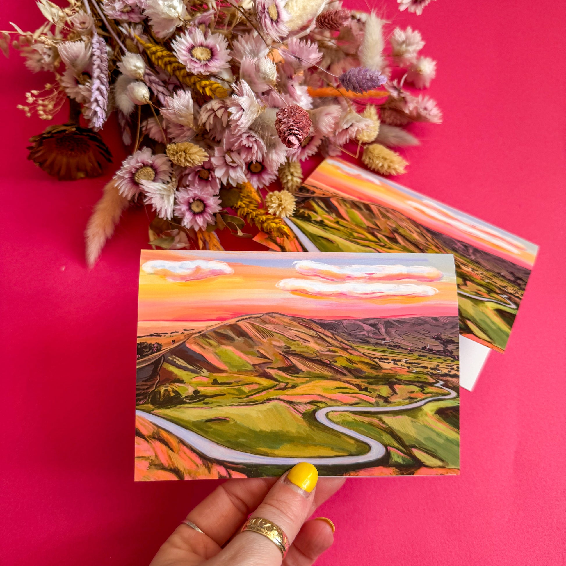 Hand holding a scenic postcard with a pink floral arrangement on a pink background