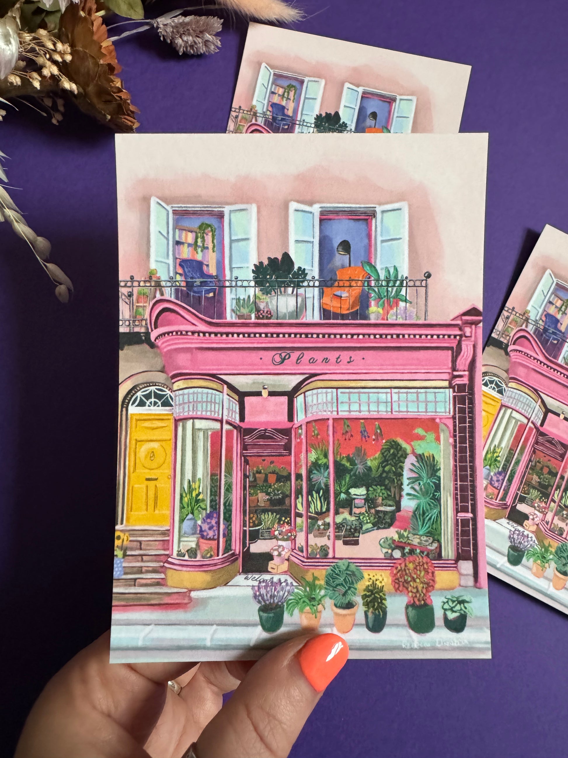 Pink shop front of a plant store with lots of greenery inside, a balcony above with windows showing chairs and a bookcase and more flowers. A yellow door to the side with a sunflower vase resting on the steps by Rebecca Elizabeth Draws. 