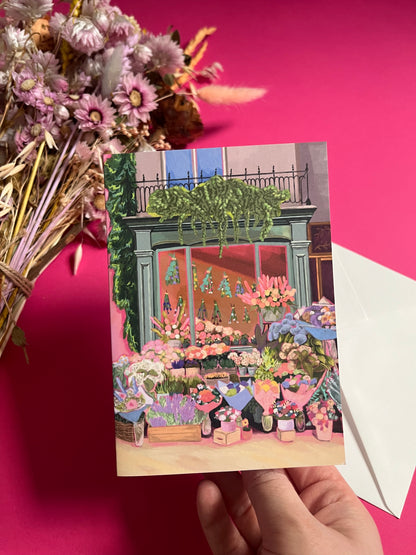 Greeting card of a beautiful flower shop, the window shows the flowers drying inside and outside there are rows of bunches of flowers and pots of plants. Soft brown and pink with bright highlights throughout. With a pink back piece & logo.