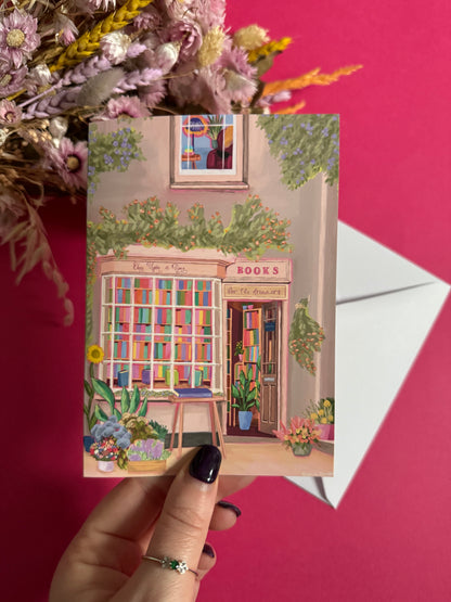 An illustration of a cute whimsical bookshop, the window shows the bookshelves inside full of bright coloured book spines with lots of flowers. The back of the card is pink with the Rebecca Elizabeth Draws logo. 