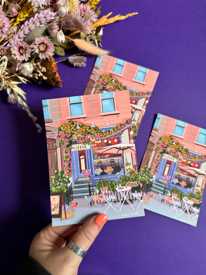 A coffee shop illustration with a blue exterior, an outside table and umbrella and building windows surrounding. This illustration is filled with lots of plants and flowers and features cups of coffee and baked goods. Illustrated by Rebecca Elizabeth Draws. 